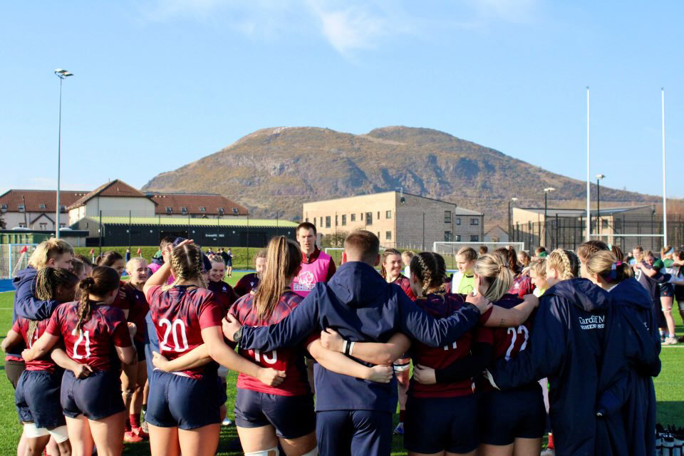 A group of women's rugby players stand in a circle on a training ground. In the background is a tall hill.